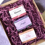 A box filled with purple shredded paper containing three rectangular soap bars labeled "Queen," "Angel," and "Wild Flower."